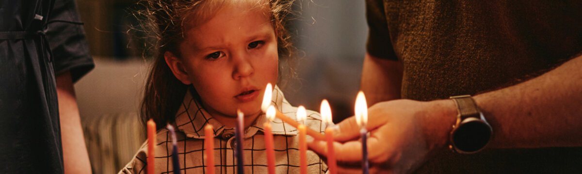 Caucasian child watching adult light Hanukkah menorah candles celebrating Jewish tradition with family. Visual ideal for holiday marketing, cultural awareness campaigns, close up