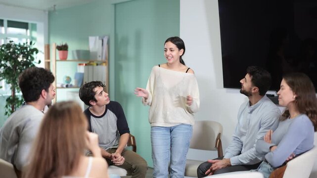 Young woman stands in front of other students who are sitting in a circle and listening to her speech. Female student came to the club of foreign languages 
