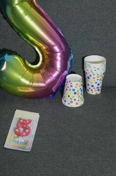 Colorful party supplies with number balloon and decorated cups on a gray couch for celebration event