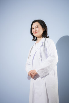 [水色背景]聴診器をつけて白衣を着る女性医師、笑顔の日本人ドクターのポートレート
