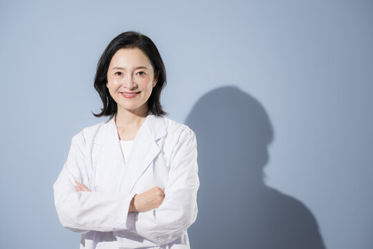 [Light blue background] A portrait of a Japanese female doctor in a white coat, arms crossed in a pensive pose, smiling and looking directly at the camera