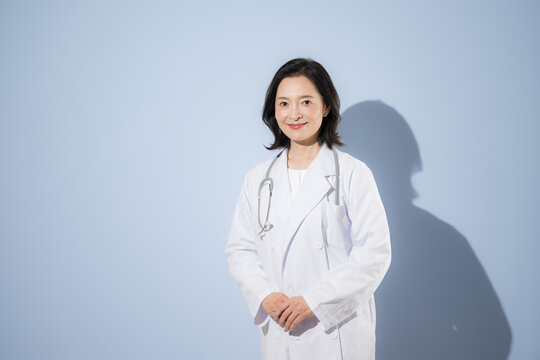 [水色背景]聴診器をつけて白衣を着る女性医師、笑顔でカメラ目線日本人ドクターのポートレート