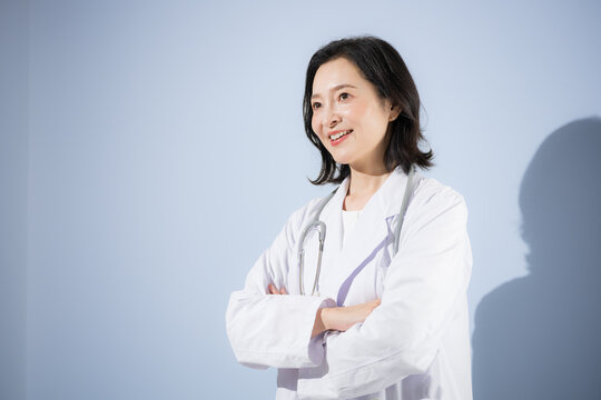 [水色背景]聴診器をつけて白衣を着て考える腕を組むポーズをする女性医師、笑顔の日本人ドクターのポートレート