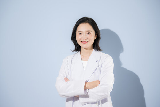 [水色背景]白衣を着て考える腕を組むポーズをする女性医師、笑顔でカメラ目線日本人ドクターのポートレート
