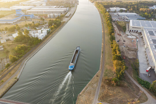 Aerial view of a cargo barge navigating the Albert Canal through an industrial area with warehouses and green surroundings in Genk, Flanders, Belgium.
