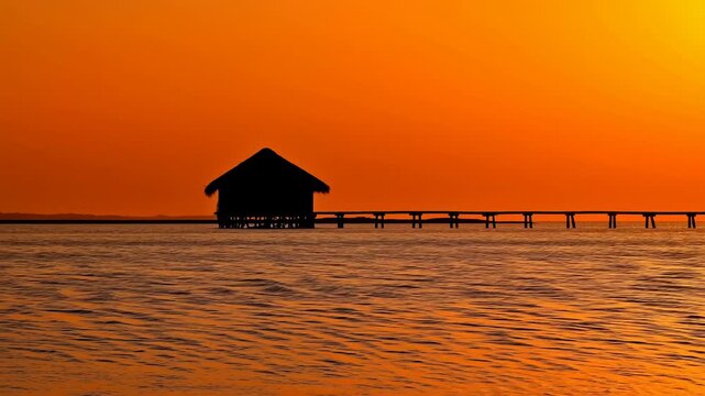 Silhouetted stilt house stands over gently rippling water beside long narrow pier, framed by glowing orange sky and warm reflections stretching across calm horizon line