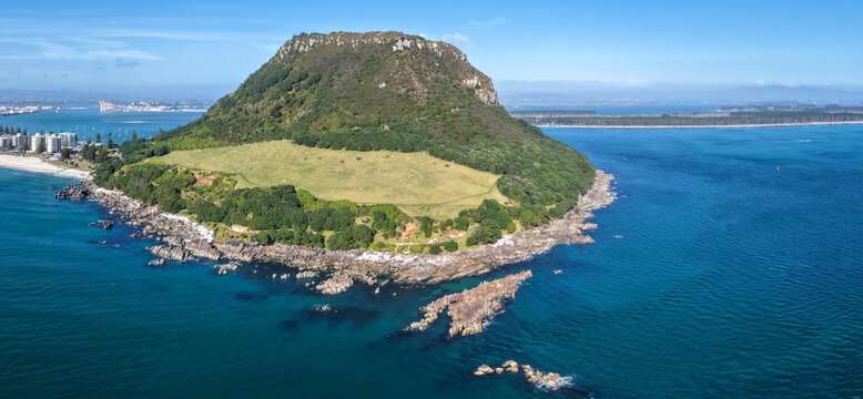 Aerial view of Mount Maunganui, a prominent volcanic cone surrounded by blue ocean waters, rocky coastline, and green slopes in Tauranga, Bay of Plenty Region, New Zealand.