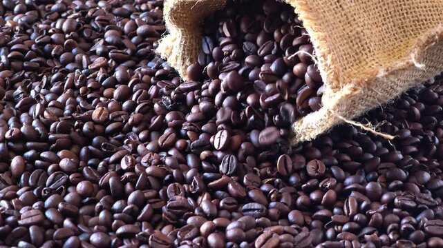 Close up of a burlap sack spilling aromatic roasted brown coffee beans over a dense background of freshly processed coffee.