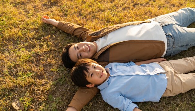 Lying father-son resting in park on patchy grass wearing brown jacket and light blue shirt
