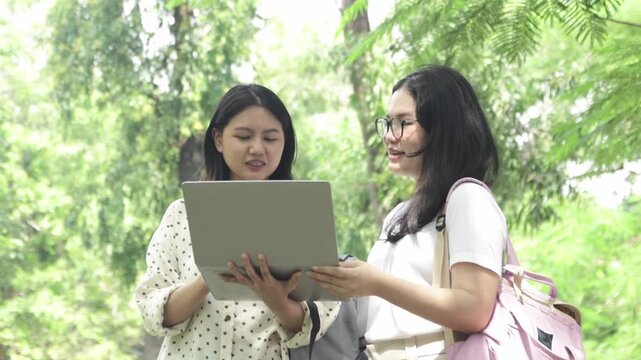 Two knowledgeable and self assured Asian female students stand on a university walkway amidst nature wearing backpacks carrying laptops review lessons together while conversing warm genuine smiles.