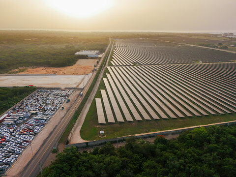 Aerial view of a large solar farm with rows of photovoltaic panels, a road with a parking lot, and greenery at sunset in San Pedro de Macoris, San Pedro de Macoris, Dominican Republic.