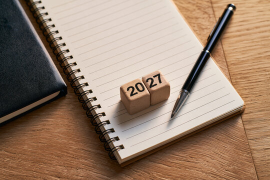 Wooden blocks displaying the year 2027 resting on an open notebook with a pen on a wooden desk.