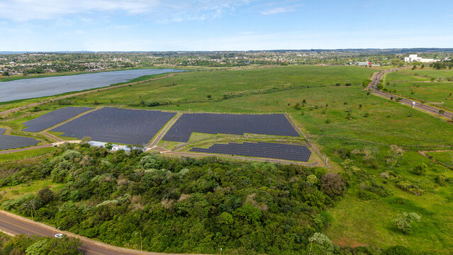 Aerial view of a large solar farm with rows of photovoltaic panels situated in a green landscape near a river and the city of Posadas, Misiones Province, Argentina.