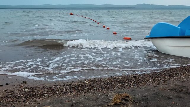 Pebbled shore of a lake or sea with gentle waves lapping against the beach. Small orange buoys line a swimming boundary extending into the water.