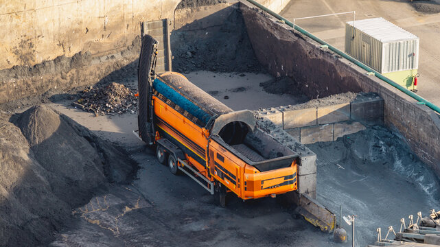 Aerial view of a mobile trommel screening machine operating among piles of dark granular material inside an industrial processing yard in Brescia, Lombardy, Italy.