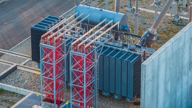 Aerial view of a high-voltage transformer in a power substation, featuring red insulators, metal busbars, and electrical components in Trino, Piedmont, Italy.