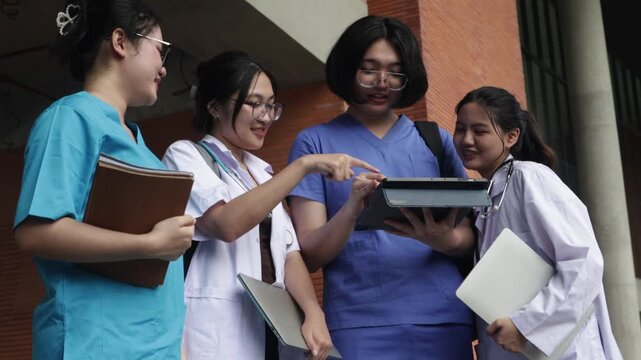 Group asian medical students dressed in blue scrubs and white coats walk and converse with one another on the campus of a modern university.

