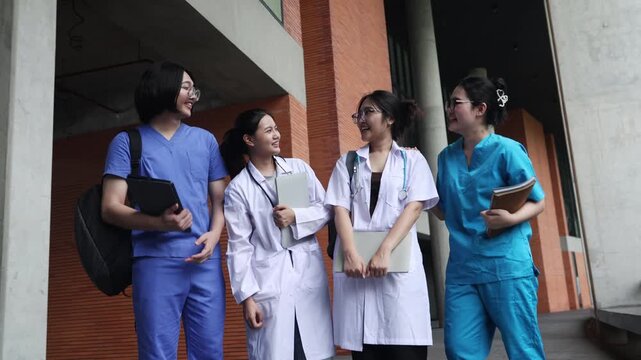 Group asian medical students dressed in blue scrubs and white coats walk and converse with one another on the campus of a modern university.
