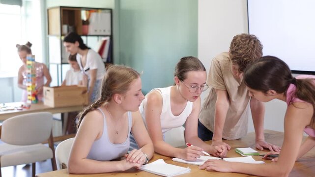 Group of teenage boys and girls sit at table, talking and writing notes in notebooks while collaborating on school assignment 