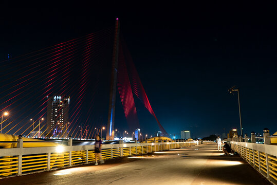Illuminated Tran Thi Ly Bridge at night in Da Nang, Vietnam, photographed from the pedestrian walkway with city lights and people enjoying the evening atmosphere.