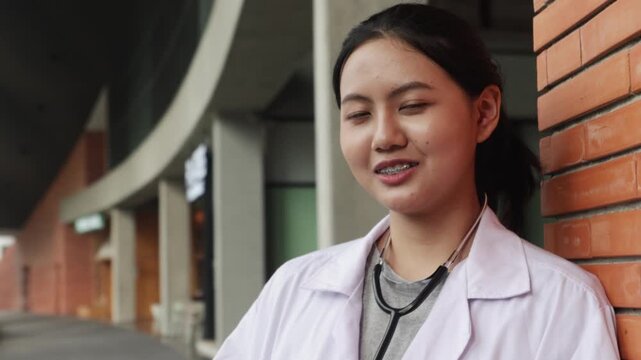 Portrait lovely Asian female medical student smiling at the camera she is wearing medical uniform holding laptop inside university building studying at an institution renowned excellent teaching.
