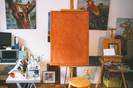 A large orange primed canvas rests on a wooden easel inside a bright vacant art studio. Authentic local small business concept for creative art classes.
