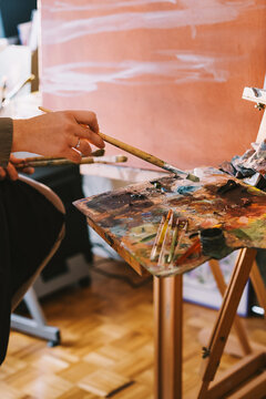 A wooden paint palette sits on a stand while a female artist paints in her art studio. Authentic local small business and art education concept.