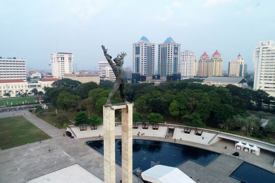 Aerial view of the Liberation of West Irian Monument in Lapangan Banteng Park surrounded by lush trees, a reflecting pool, and modern city buildings in Sawah Besar, Jakarta, Indonesia.