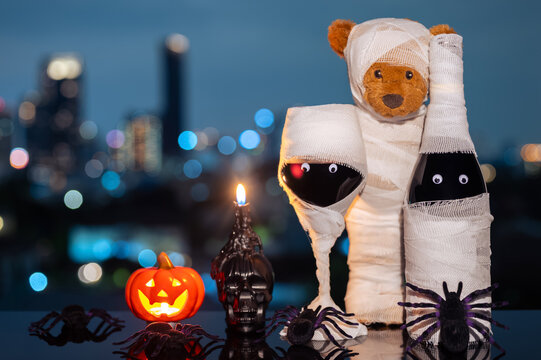 Halloween mummy bear, wine bottle, and glass with pumpkins and skull candle against a city skyline bokeh at night.