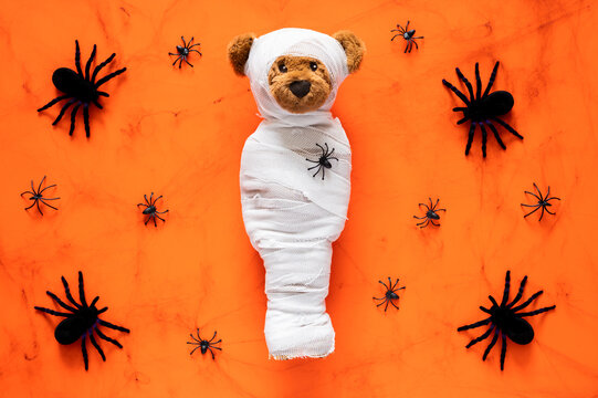 Halloween teddy bear mummy wrapped in gauze with black spiders on orange background.