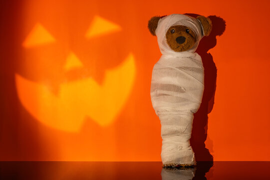 Halloween mummy teddy bear standing against an orange wall with a jack o lantern face shadow.
