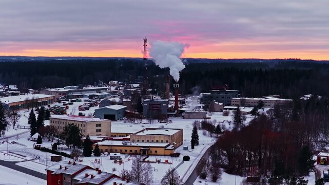 Aerial view of an industrial facility with a smoking chimney amidst a snow-covered landscape under a vibrant sunset sky in Latvia.
