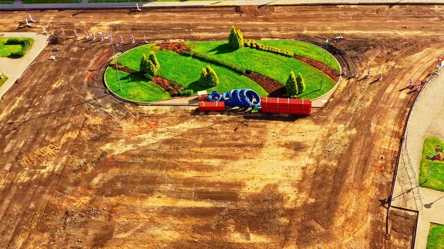 Aerial view of a construction site with a landscaped roundabout, green grass, small trees, bare earth, and construction materials including blue pipes and red bundles in Latvia.