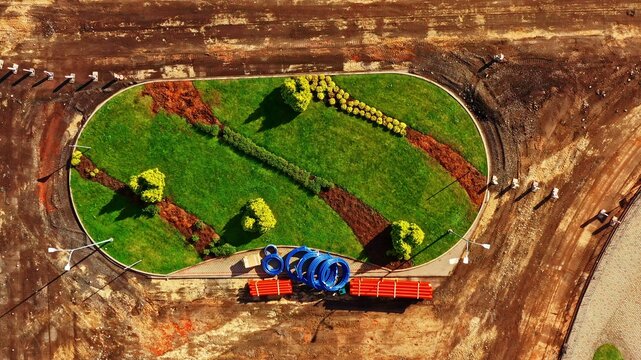 Aerial view of a landscaped oval roundabout under construction with green grass, small trees, blue pipes, and orange pipes in Latvia.