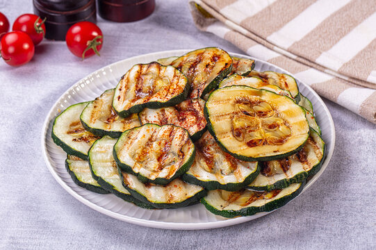Grilled zucchini slices with charred marks and spice seasoning on white plate, cherry tomatoes and striped napkin in background