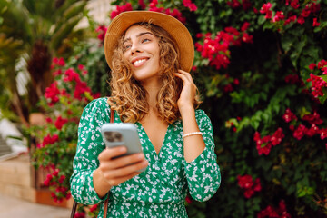 Woman in hat and dress stands outdoors among bright flowers, holding phone. Smiling woman looks at phone against backdrop of flowering bushes at sunset. Concept of technology, leisure. Lifestyle. © Koval988