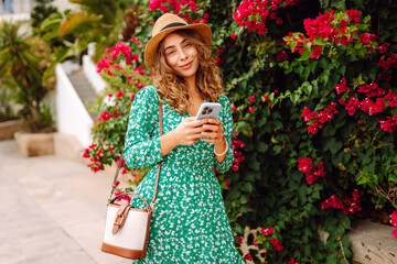 Woman in hat and dress stands outdoors among bright flowers, holding phone. Smiling woman looks at phone against backdrop of flowering bushes at sunset. Concept of technology, leisure. Lifestyle. © Koval988