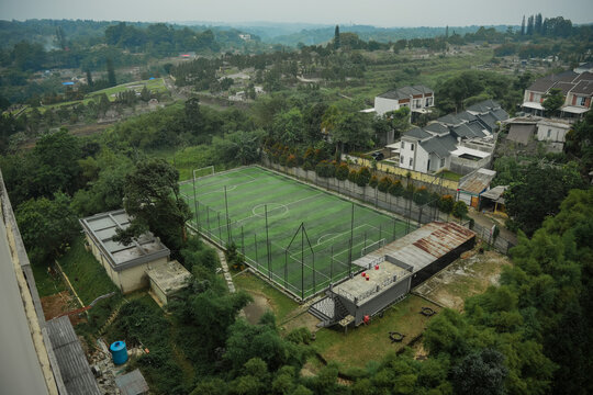 Aerial view of a mini soccer field surrounded by lush green trees and residential houses in Bogor, West Java, Indonesia.