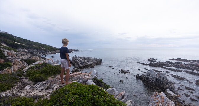 lone boy looking out at stormy late afternoon light on rocky coastline of DeKelders, South Africa