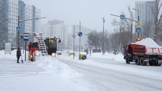 Snow removal trucks and blower operating on snowy city street beside bundled pedestrian at slushy intersection, with apartment blocks, bare trees, traffic signs, and winter fog