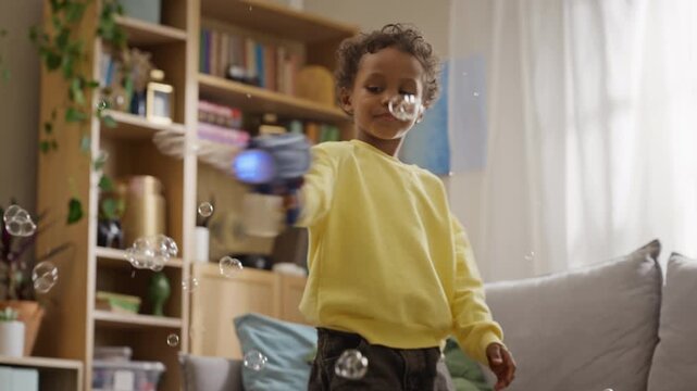African American boy shooting soap bubbles with automatic toy blower indoors in slow motion. Footage suited for toy advertising, parenting blogs, and family lifestyle marketing campaigns