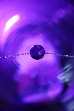 Close-up of a purple gemstone on a silver chain, set against a blurred purple background. The gemstone is round and shiny, reflecting light.