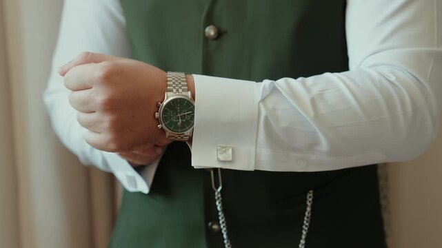 Groom preparing for wedding, adjusting cufflink on white shirt sleeve, wearing a watch and green vest. Concept of elegance