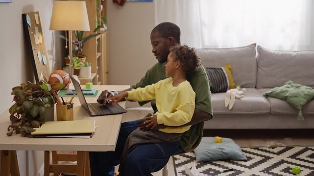 African American father typing on laptop with young son sitting on lap at home office. Ideal for marketing remote working, family tech solutions, or digital parenting