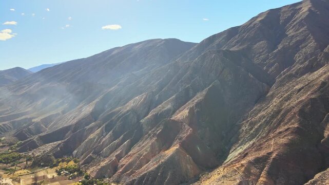 Steep colored slopes of Cerro General San Mart&iacute;n Paleta del Pintor rise above farms near Maimar&aacute; under clear daylight, seen from a moving drone.