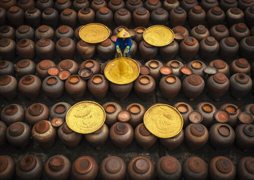 Aerial view of a worker wearing a conical hat sorting yellow grains on trays amidst rows of large ceramic jars at a soy sauce factory in Ban Yen Nhan, Vietnam.
