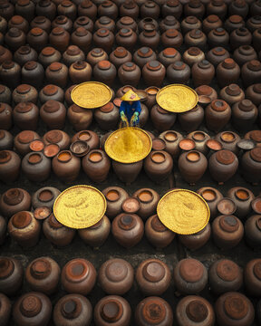 Aerial view of a worker wearing a conical hat processing soy sauce in yellow trays surrounded by numerous brown ceramic jars at a soy sauce factory in Ban Yen Nhan, Vietnam.