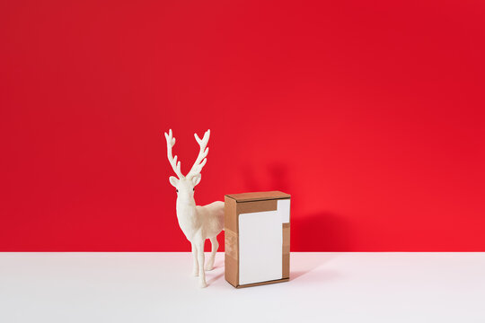 Christmas delivery concept with cardboard package and white reindeer figurine on red background