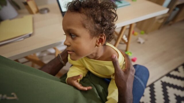 POV of African American toddler boy looking up into camera while embracing father at home desk. Footage suits advertising for family insurance, modern fatherhood, and work life balance