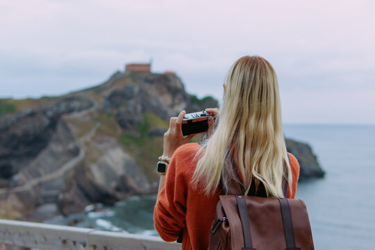 Woman with blonde hair and backpack capturing the iconic landscape of gaztelugatxe island with her camera, exploring the historic spanish coast